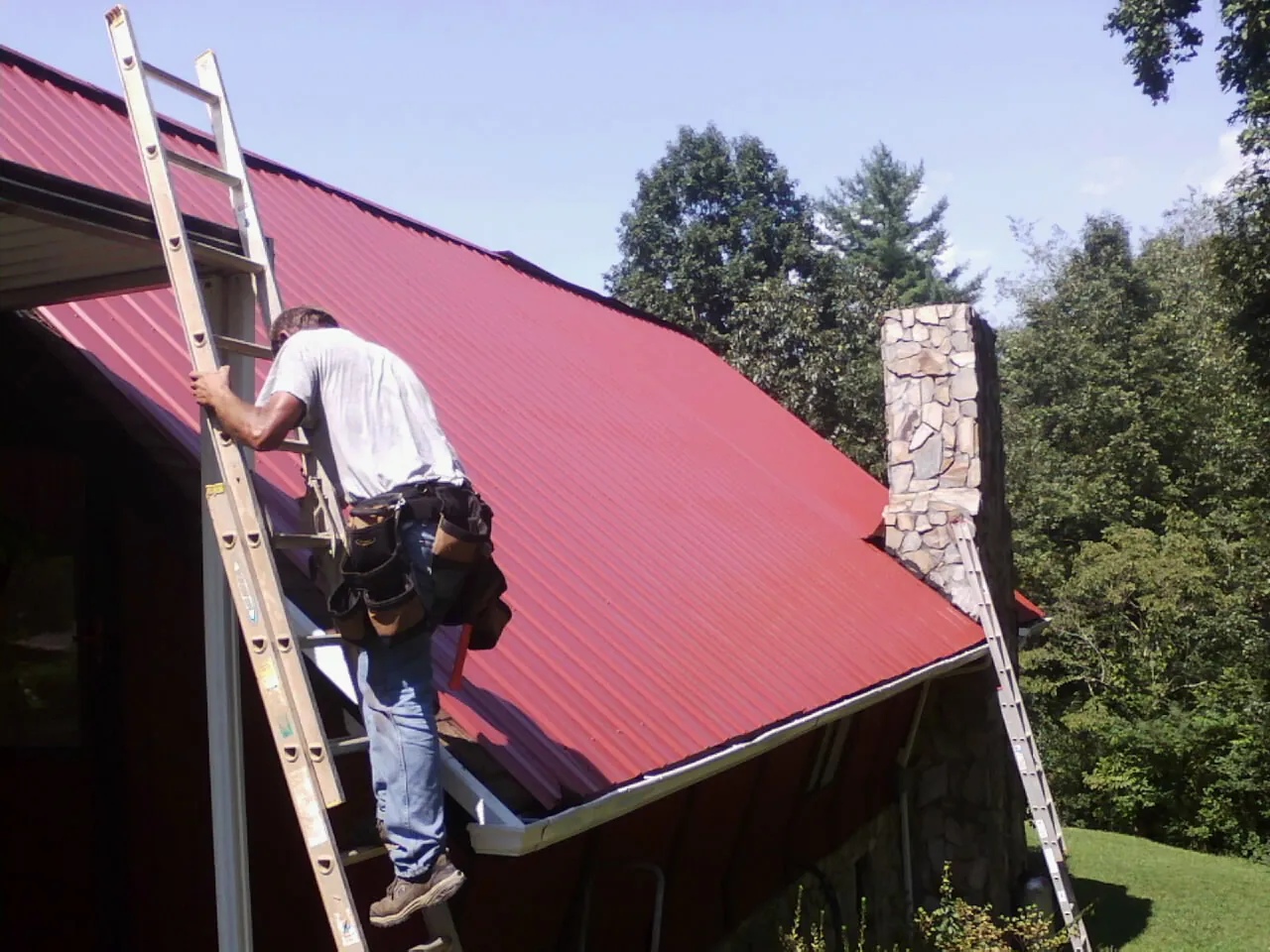 Roofer on a ladder beside a red metal roof and stone chimney — Soffit & Fascia Repair by Roofing Brothers in Duarte